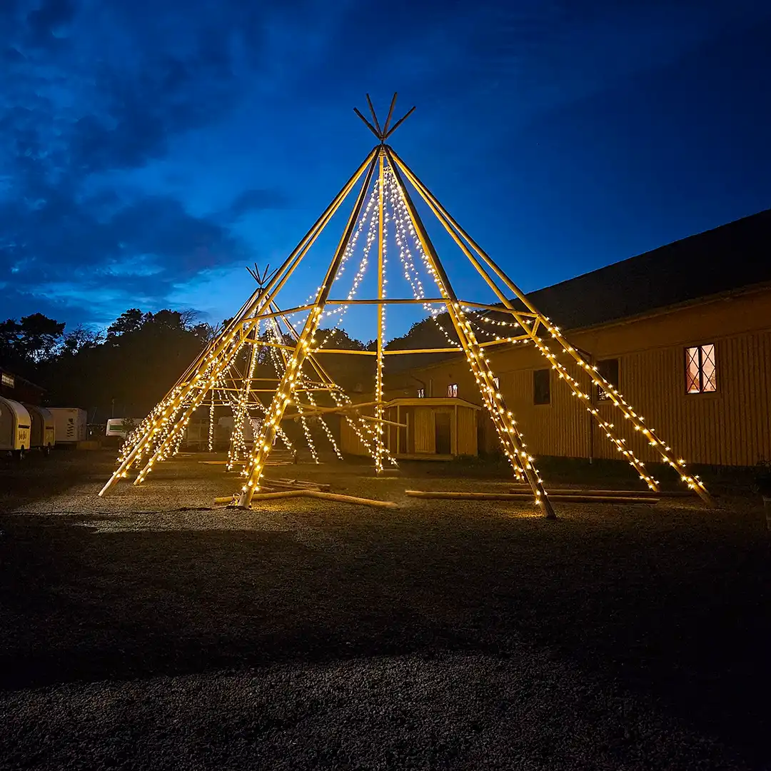 Oplyst tipi-stel med lyskæder i en gårdsplads om aftenen. Perfekt til udendørs events, bryllupper og firmafester i naturskønne omgivelser.