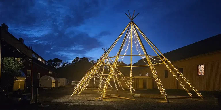 Oplyst tipi-stel med lyskæder i en gårdsplads om aftenen. Perfekt til udendørs events, bryllupper og firmafester i naturskønne omgivelser.