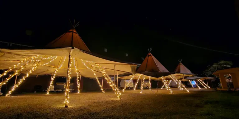 Stor tipi med åbne sider fyldt med mennesker, der nyder et udendørs arrangement i solen.