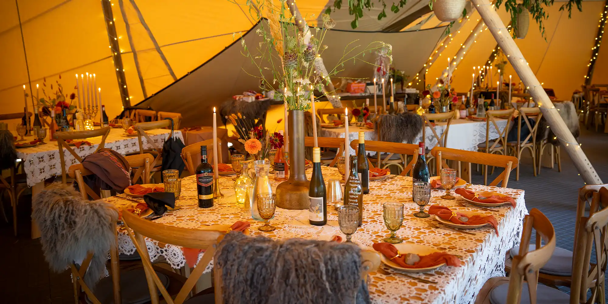 Elegant dækket festbord med vin, blomster og stearinlys i en tipi med hyggelig belysning – perfekt til bryllupper og eksklusive events.
