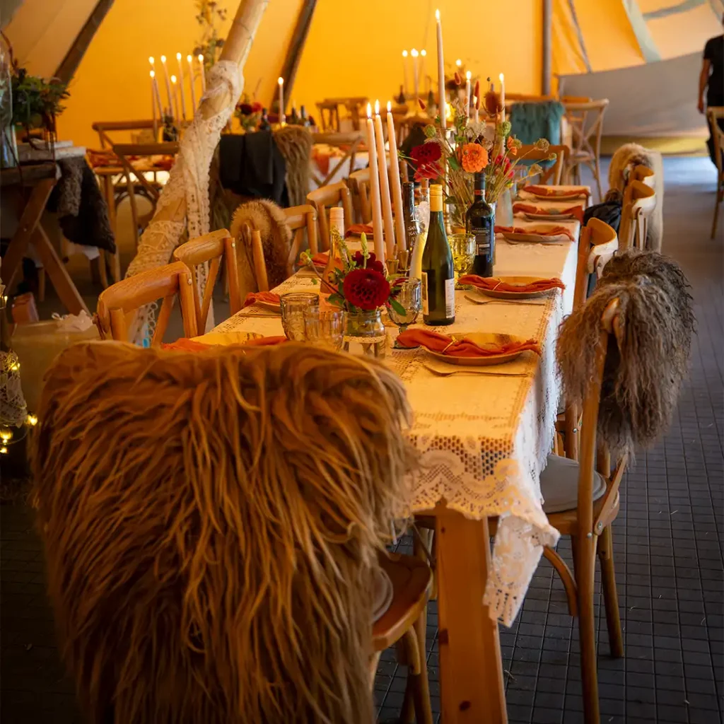 Langbord dækket med elegante duge, levende lys og rustik borddækning i en stemningsfuld tipi, ideel til bryllupper, firmafester og private arrangementer.