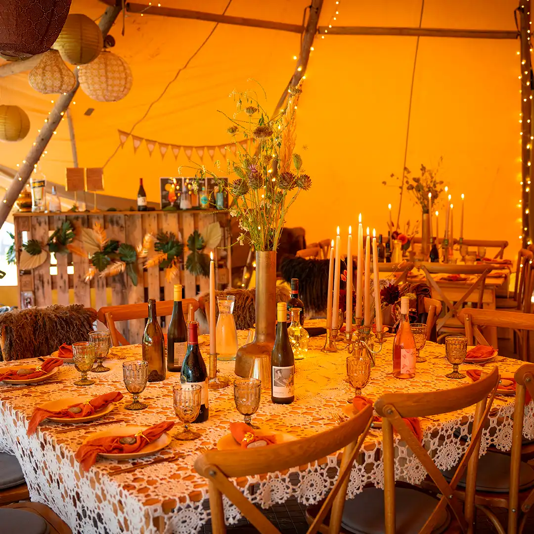 Smukt dækket festbord med stearinlys, vinflasker og blomster i en hyggelig tipi – perfekt til bryllupper og private fester.