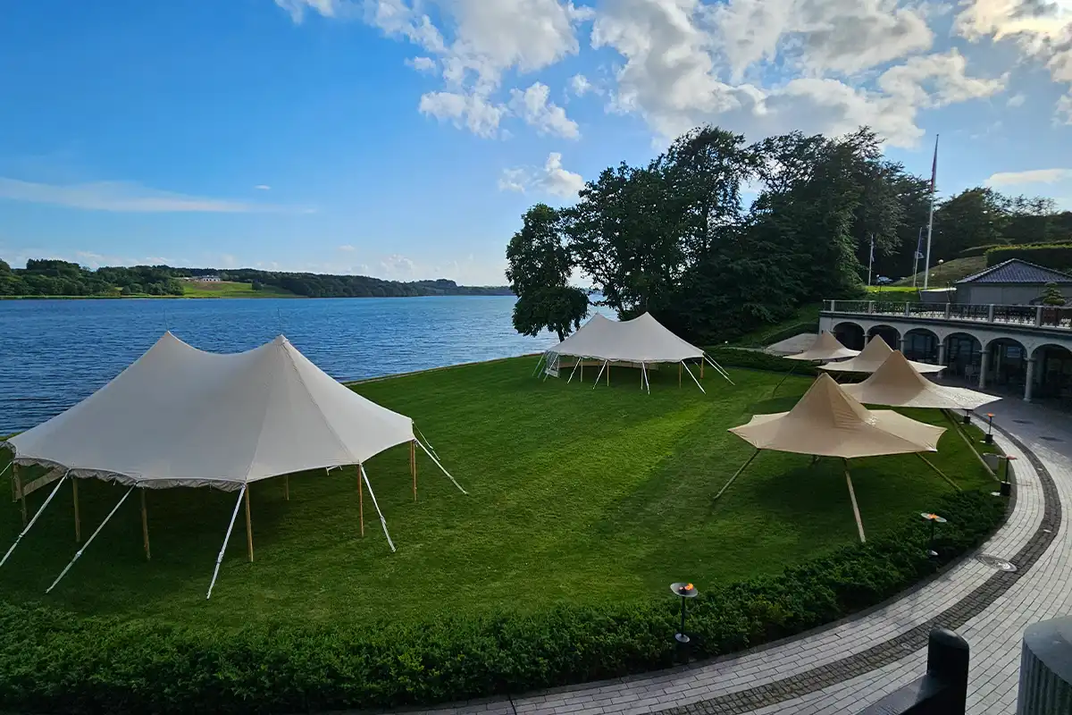 Flere mastetelte opstillet på en grøn plæne ved en smuk sø, omgivet af frodig natur og blå himmel – ideelt til bryllupper, firmaevents, sommerfester og udendørs arrangementer i naturskønne omgivelser.