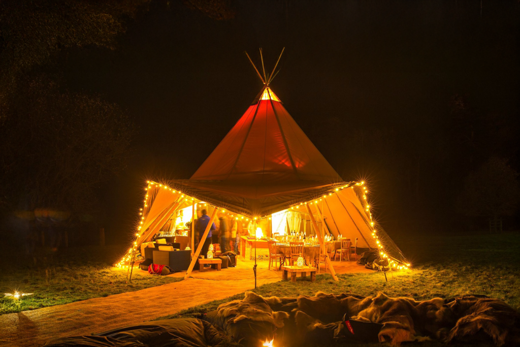 En smukt oplyst tipi i natten, dekoreret med lyskæder og omgivet af gæster, bål og hyggelige siddepladser i en naturlig udendørs atmosfære.