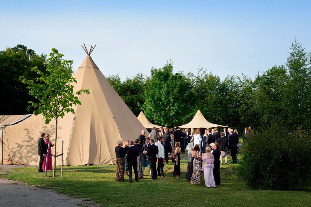 En stor tipi opsat i grønne omgivelser, hvor velklædte gæster mingler ved et elegant udendørs bryllups- eller firmaevent.