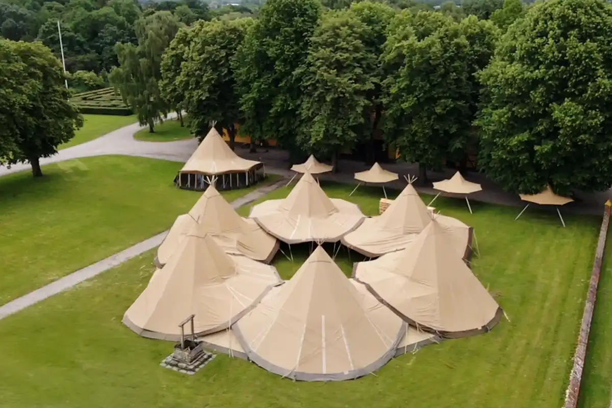 Luftfoto af en imponerende tipi-opsætning til events, bryllupper og firmafester, omgivet af grønne arealer og træer. Perfekt til store sammenkomster i naturskønne omgivelser.