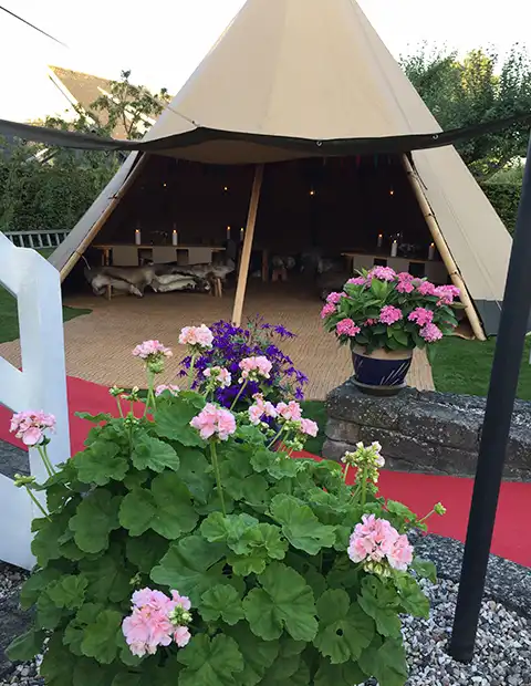 En smukt dekoreret tipi med åbning ud mod en have, omkranset af blomster og en rød løber. Indretningen byder på træborde, bænke med fåreskind og levende lys, hvilket skaber en hyggelig atmosfære til festlige begivenheder.