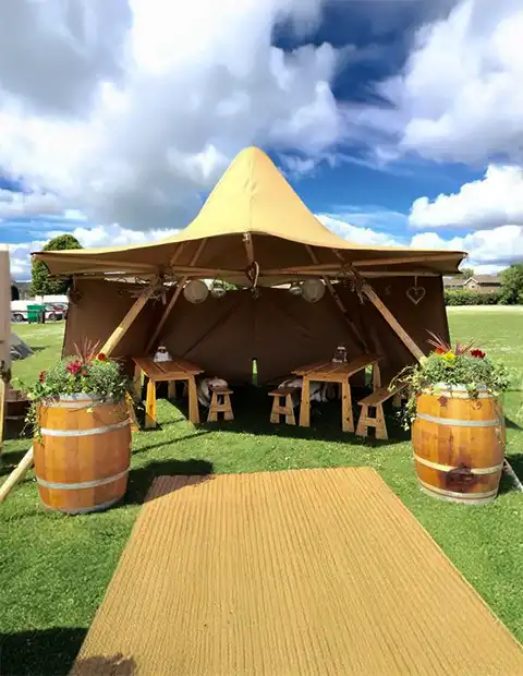 En åben tipi med en unik form, dekoreret med lanterner, blomster i vintønder og rustikke træborde med bænke. Teltet står på en grøn græsplæne under en blå himmel og er ideelt til hyggelige udendørs arrangementer og fester.