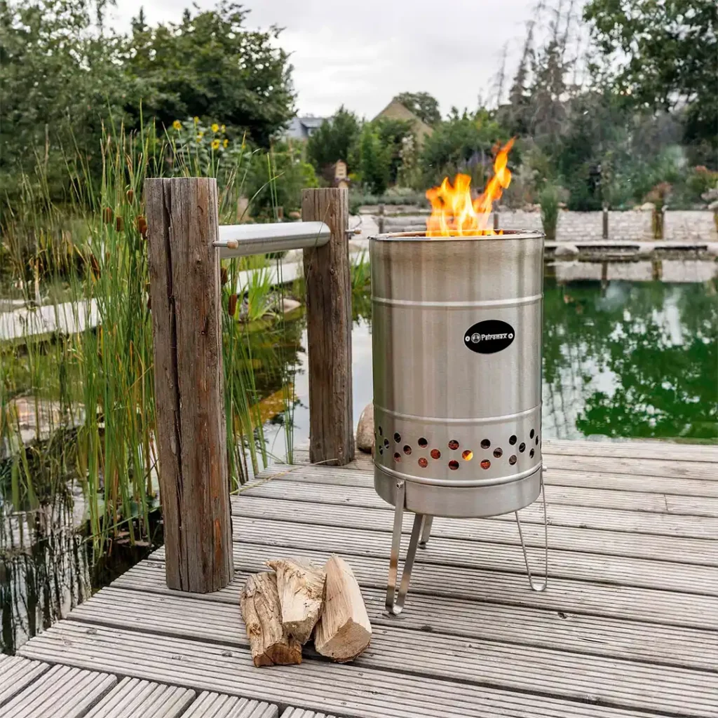 Moderne højt bålfad fra Petromax med flammer, placeret ved en idyllisk sø. Ideel til at skabe varme og hyggelig stemning ved udendørs arrangementer, fester eller firmaevents.