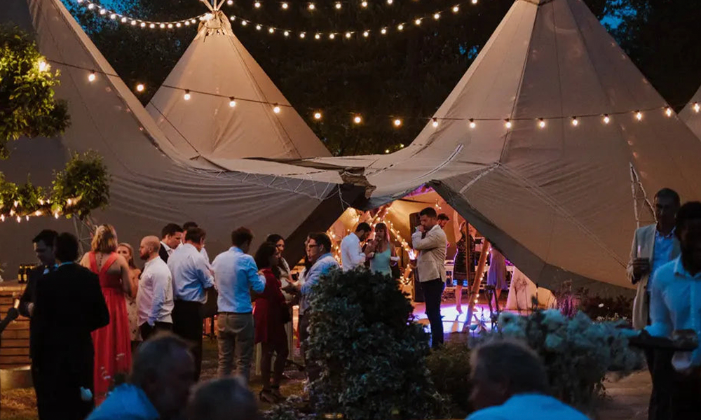 Bryllupsfest i udendørs tipi-telt dekoreret med smukke lyskæder, glade gæster, der fejrer bryllup i romantisk stemning om aftenen