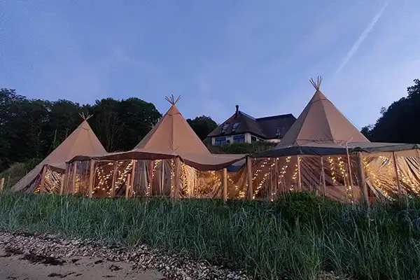 Stemningsfulde tipi-telte med lyskæder opsat ved en villa nær kysten, perfekt til bryllupper, firmafester og luksuriøse events – TipiogCo.