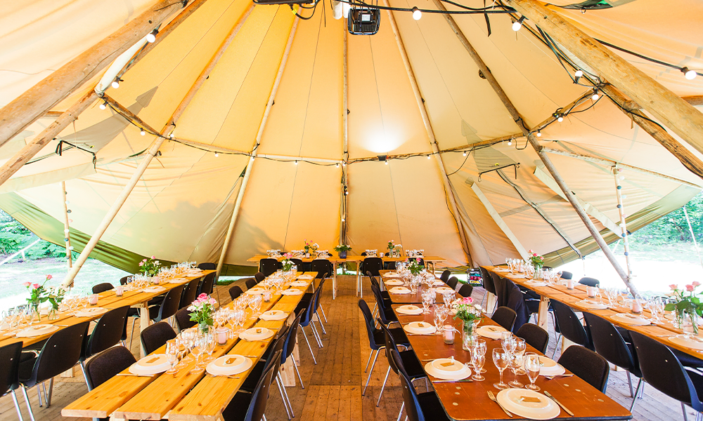 Festligt opdækkede borde i tipi-telt med blomster, glas og træborde, skaber en hyggelig og rustik atmosfære til bryllup eller fest – TipiogCo