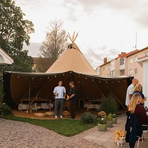 Stratus 40 tipi med åben side og ildsted i midten, set udefra gennem vindue med blomster.